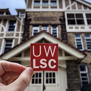 A hand holds a sticker reading, "UW LSC," in front of Hiram Smith Hall