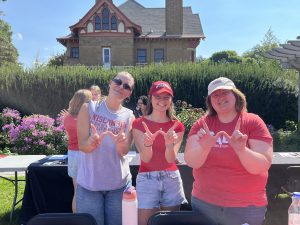 Three women, two wearing red shirts and one wearing gray, each make a "w" with their hands standing in front of a nice house in a garden. 
