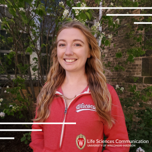A woman with pale skin, strawberry blonde hair wearing a red jacket that reads Wisconsin smiles. 